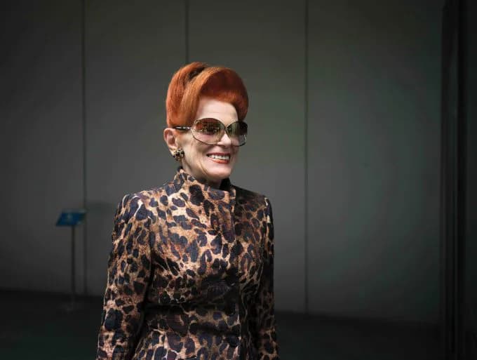 Woman with orange bob hair and sunglasses wearing a leopard-print turtleneck, posed against neutral background.