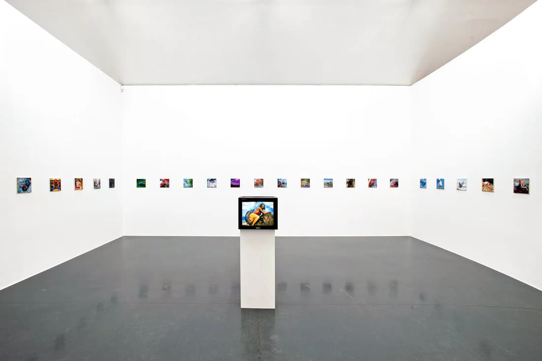 Minimalist white gallery interior with small framed artworks in horizontal line on wall and central pedestal with video monitor.