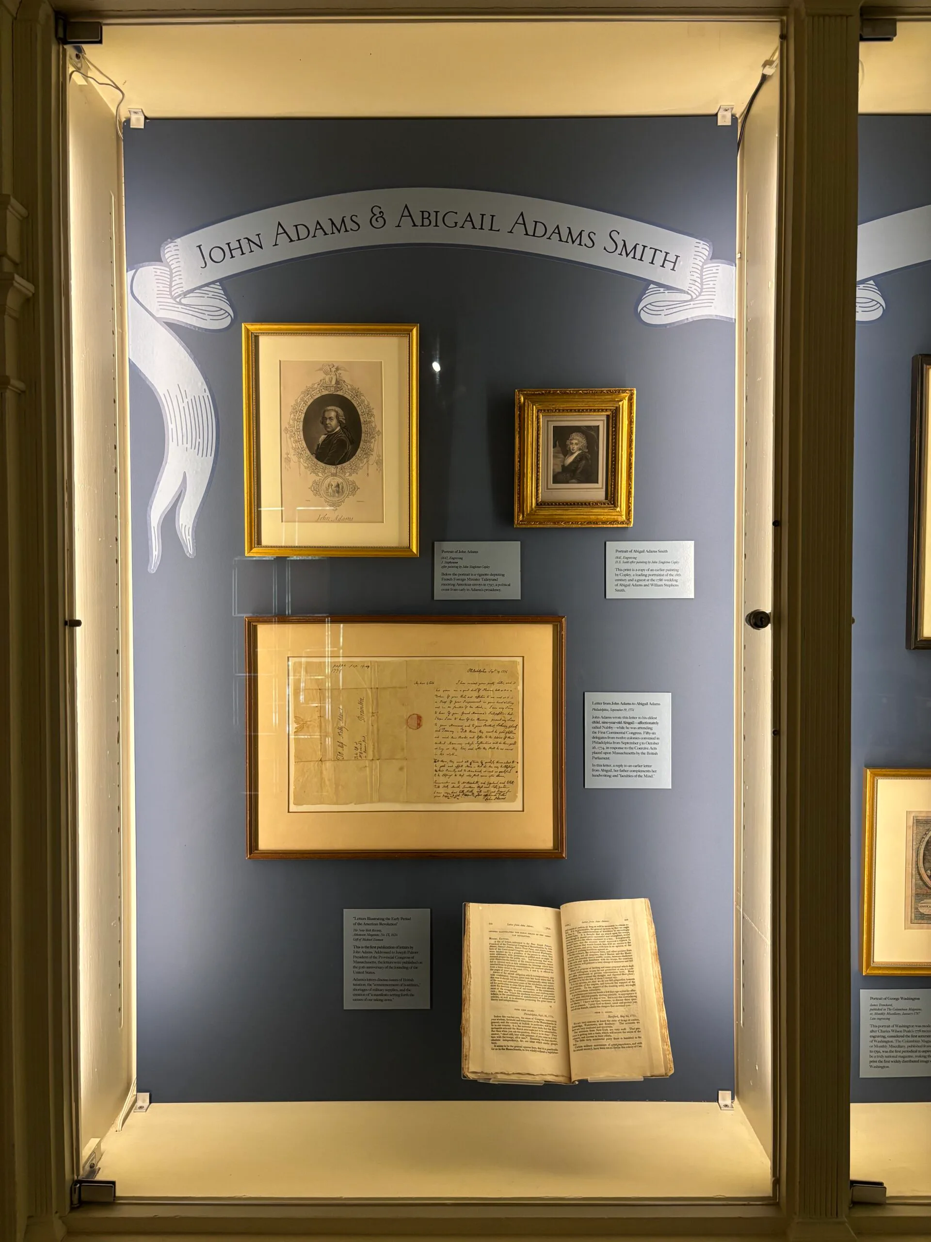 Museum display case with dark blue backing featuring framed portraits, a historical document, and an open vintage book related to John Adams and Abigail Adams Smith.