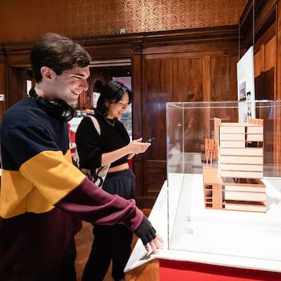 Gallery visitors examining a display case in an ornate wood-paneled interior, architectural models visible in exhibition.