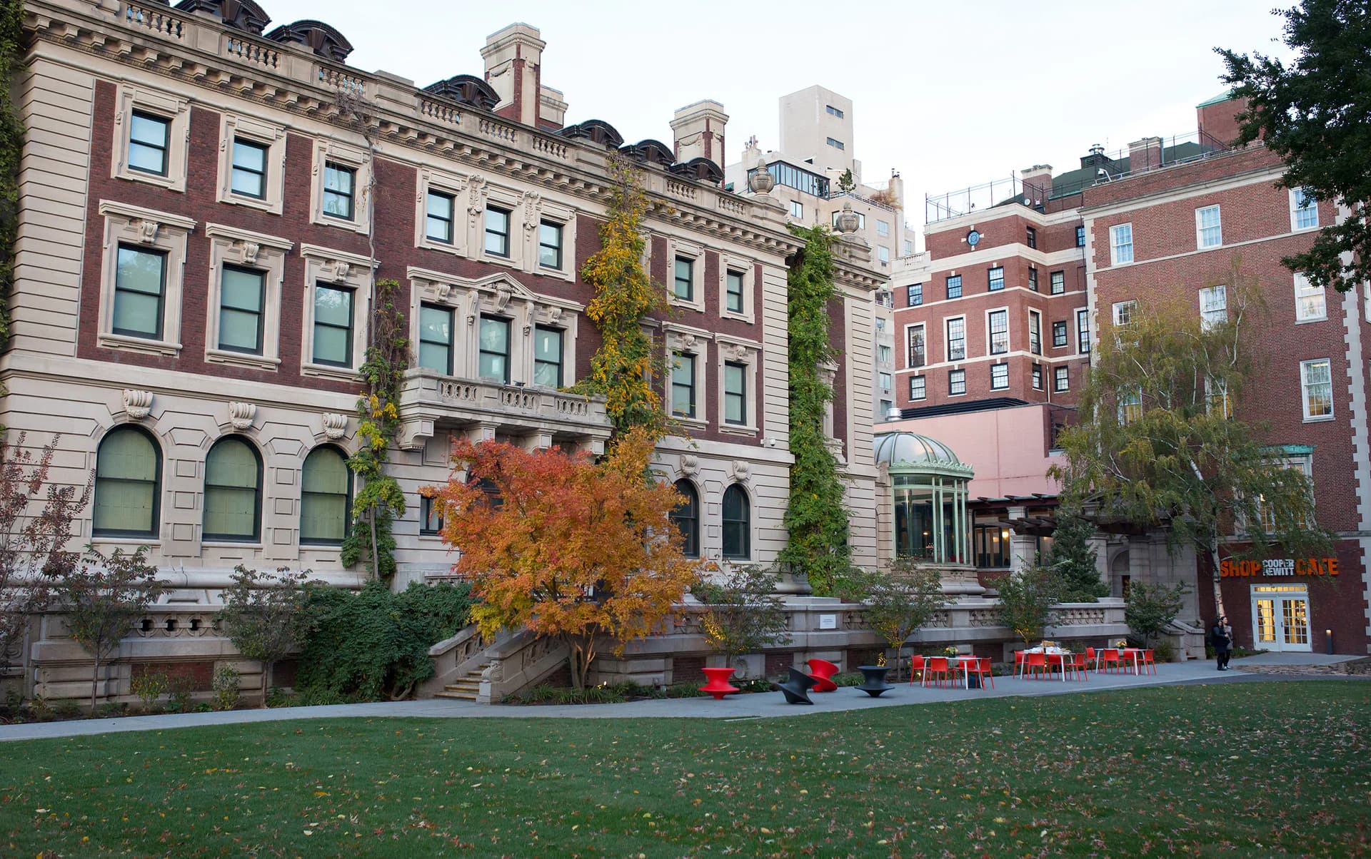 Exterior courtyard of Cooper-Hewitt museum with historic red-brick townhouses, outdoor seating, and autumn foliage.