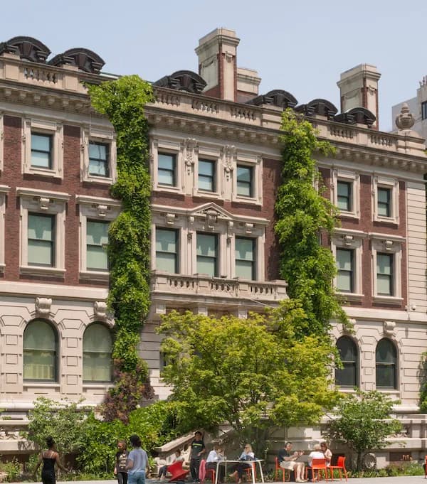 Exterior of a red-brick institutional building with classical architecture, ivy climbing the facade, and people seated at tables in a courtyard below.