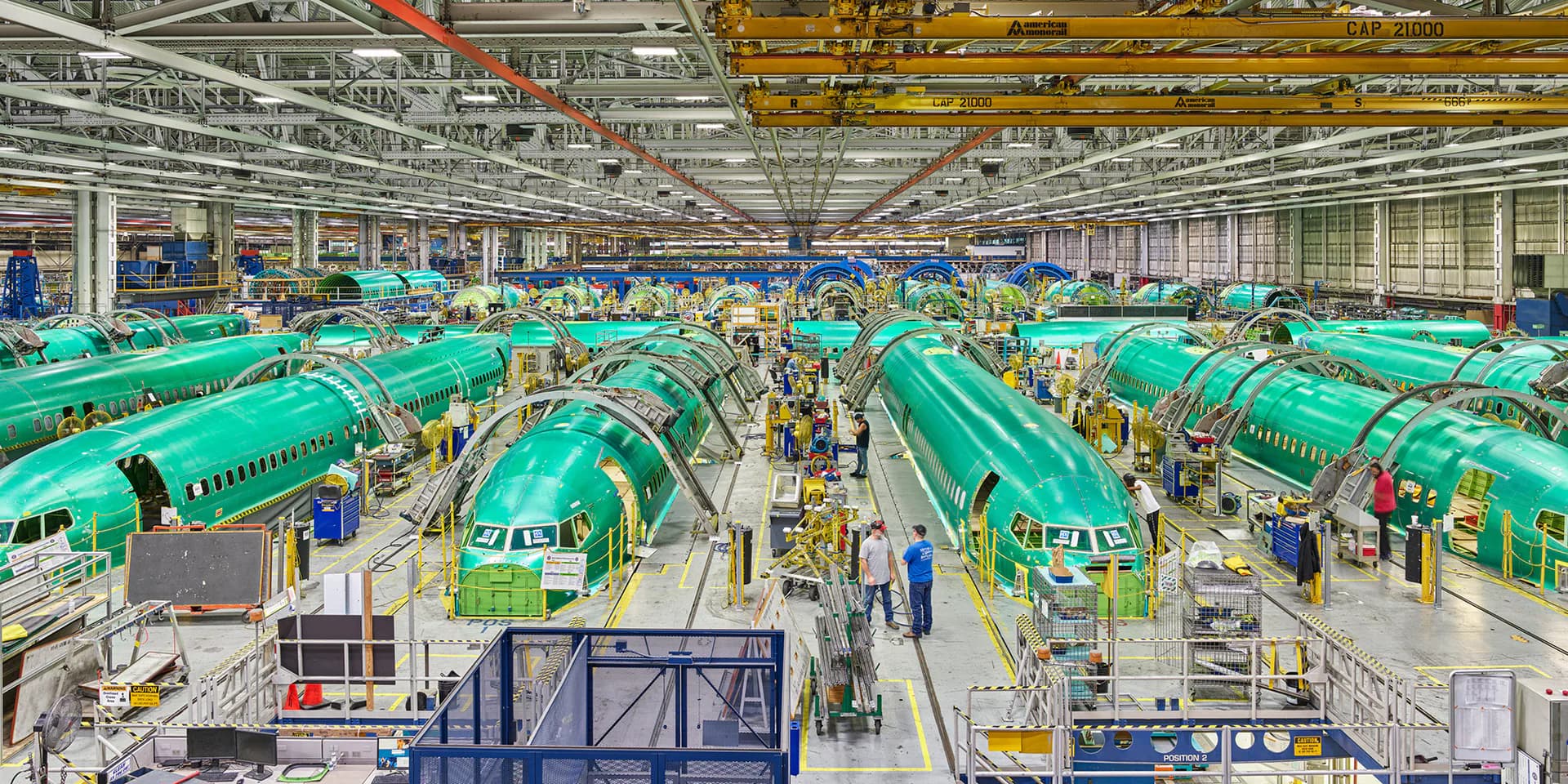 Interior of an aircraft manufacturing facility with rows of teal-green fuselages under assembly, industrial cranes overhead, and workers on the factory floor.