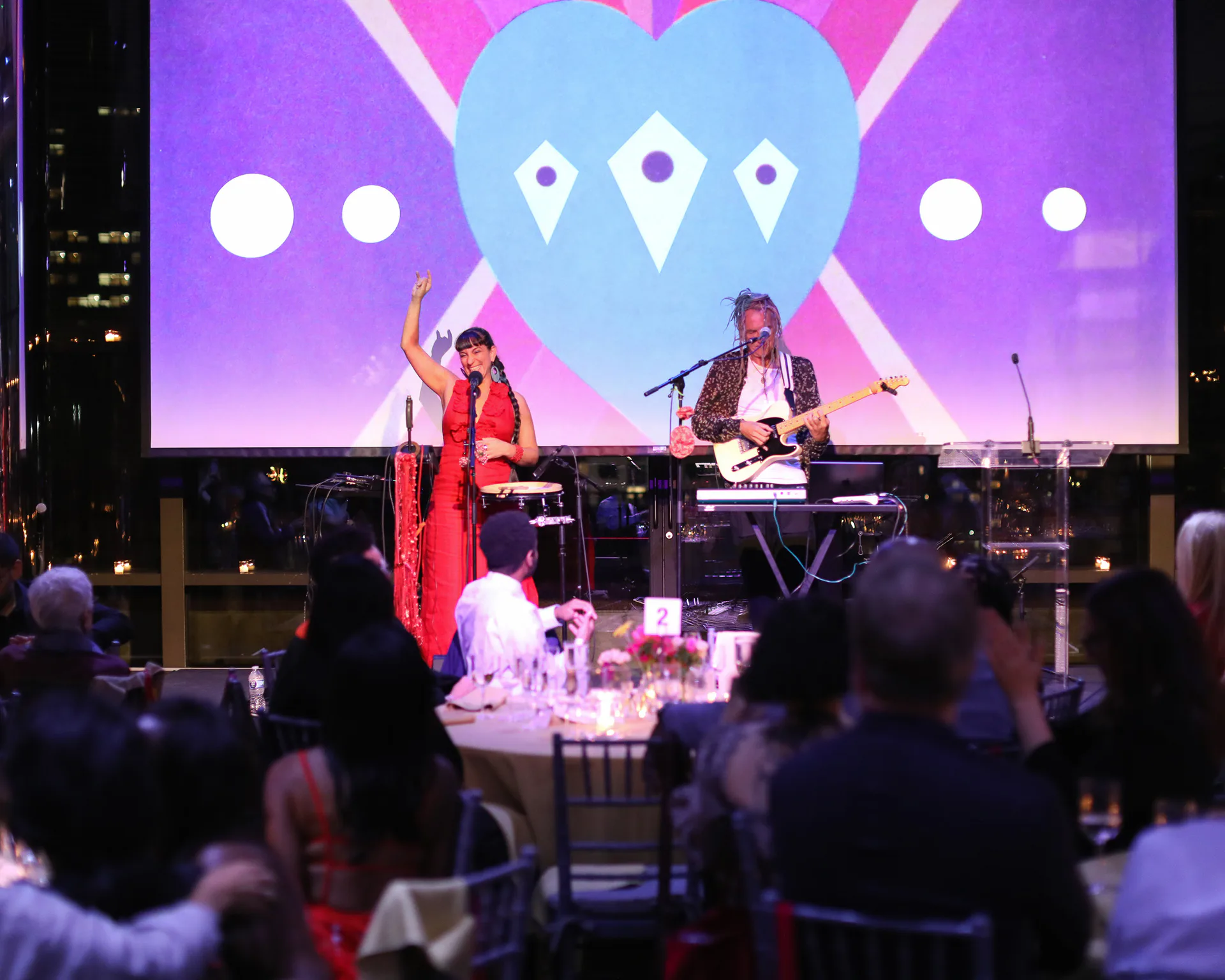 Live performance in gallery or event space with two performers on stage, one in red, against purple and blue graphic backdrop with geometric shapes; audience visible in foreground.