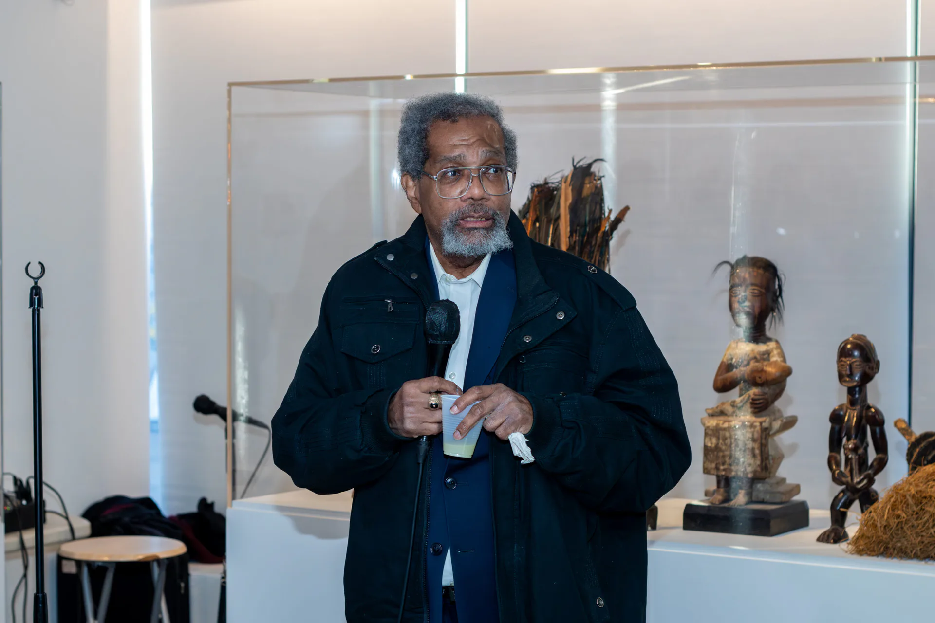 Man speaking at microphone in front of African wooden sculptures displayed in gallery cases.