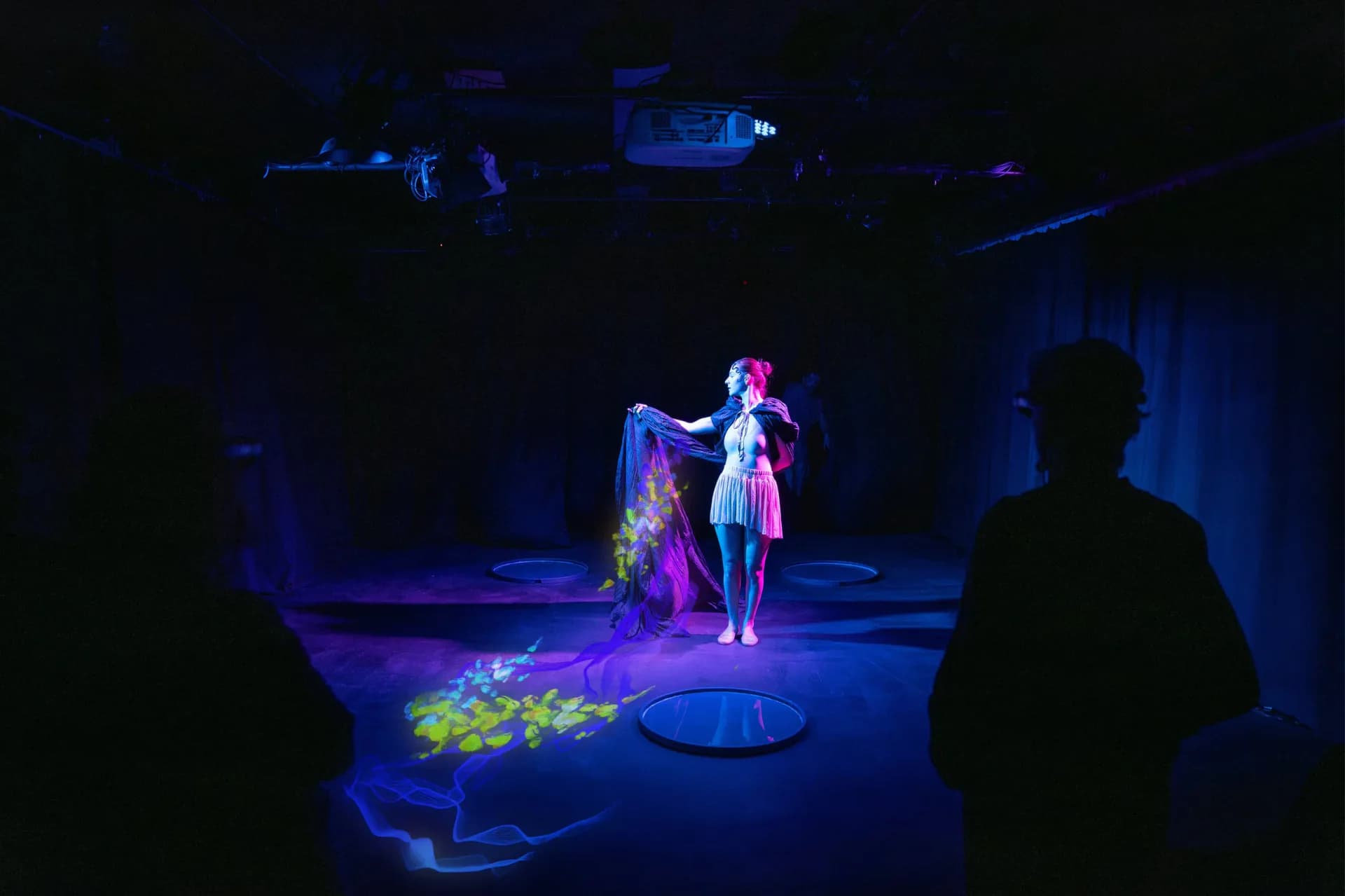 Dark theater or performance space at Culturehub with silhouetted audience member watching an illuminated performer in colorful costume under blue stage lighting.