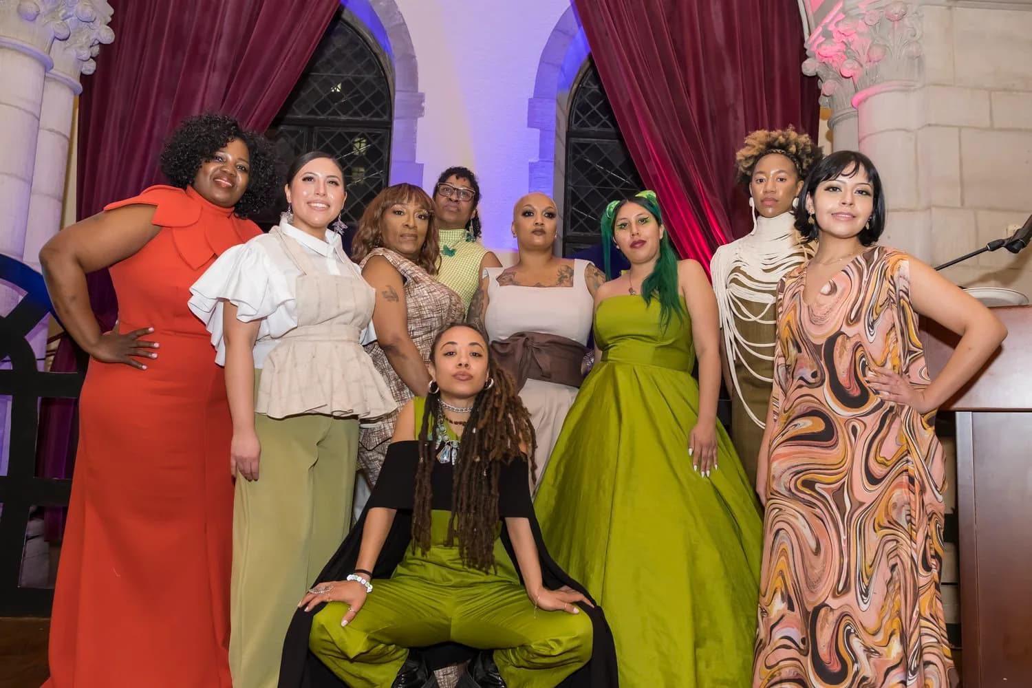 Group of people in colorful clothing posed in an elegant interior space with burgundy curtains, arched windows, and staged lighting.