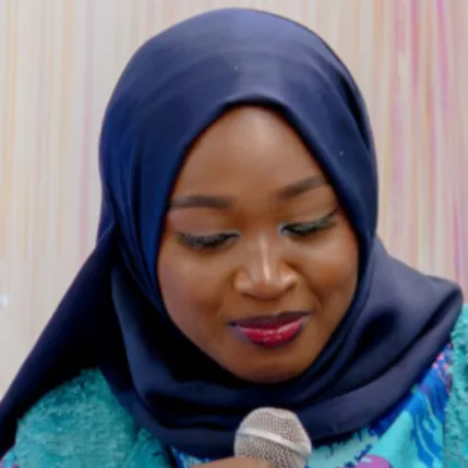 Portrait of a person wearing a navy blue hijab and holding a microphone, with warm makeup and a calm expression.