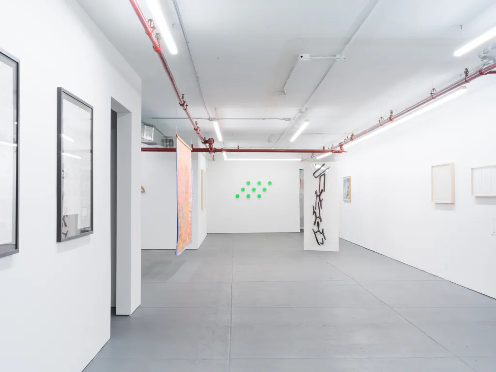 Minimalist gallery interior at D. D. D. D. with white walls, red industrial pipes overhead, concrete floor, and scattered artworks including green dots and ink drawings.