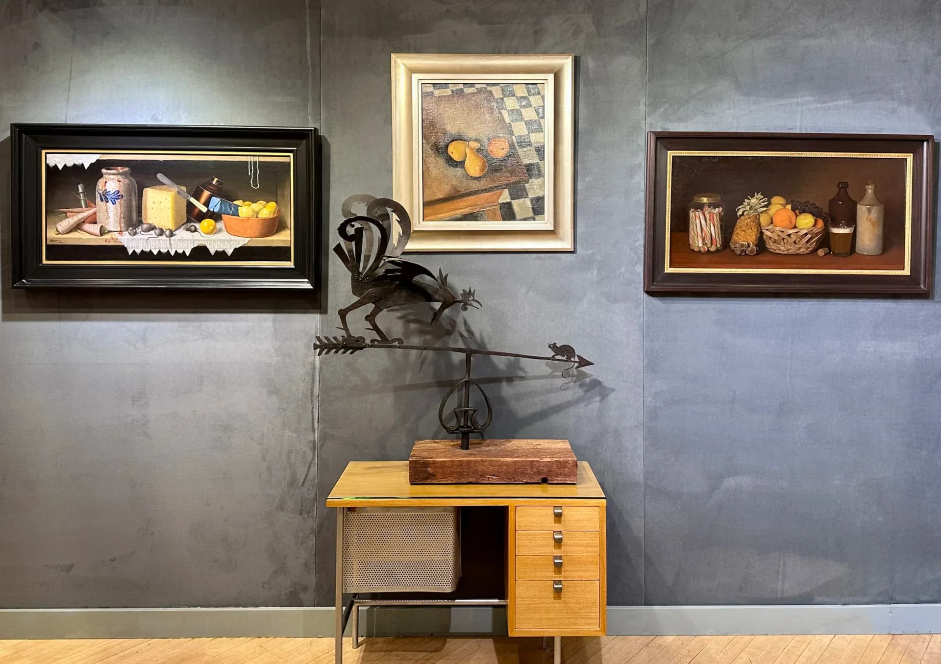 Gallery wall at D. Wigmore Fine Art displaying three framed still-life paintings above a mid-century wooden desk with sculptural bronze figure.