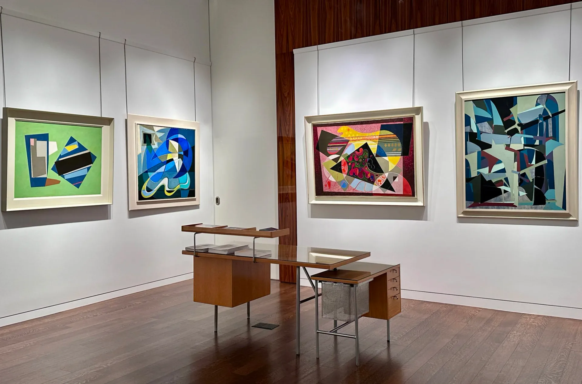 Modern gallery interior with four abstract paintings in varied color palettes hung on white walls, wooden display tables in center floor.