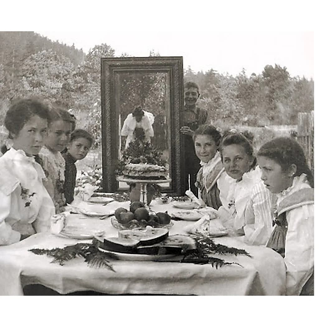 Black-and-white photograph of women in white dresses seated at an outdoor table with a mirror, forest backdrop, and mountainous landscape.