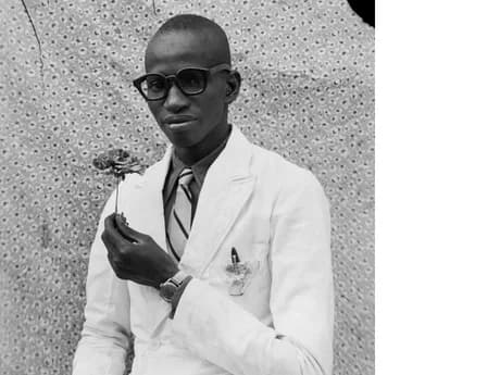 Black-and-white portrait of a man in glasses and white coat holding a small plant, against a dotted background.