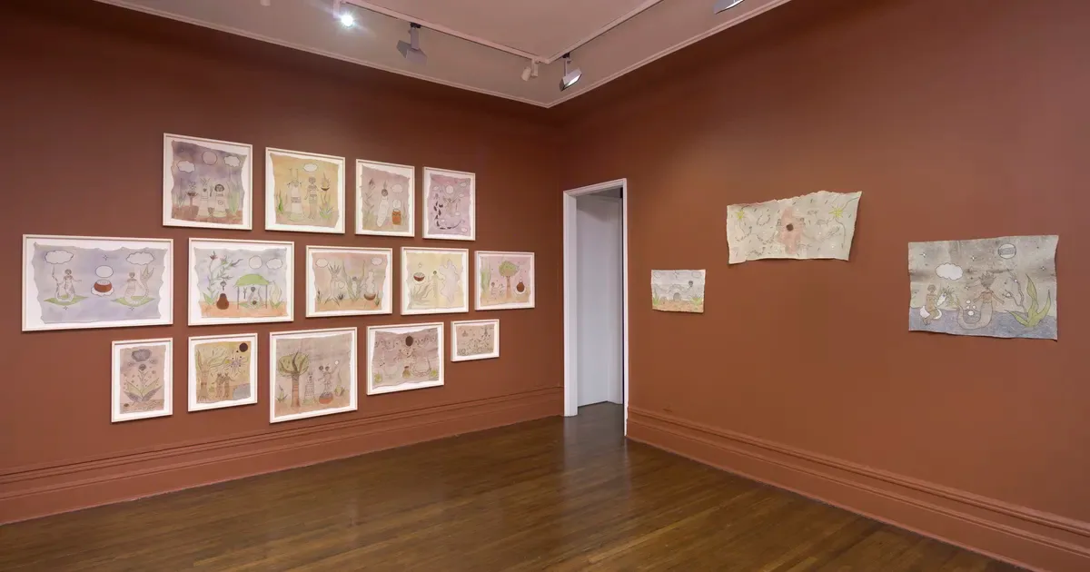 Gallery interior with terracotta-colored walls displaying framed drawings and prints arranged in salon-style groupings; wooden floor and white doorway visible.