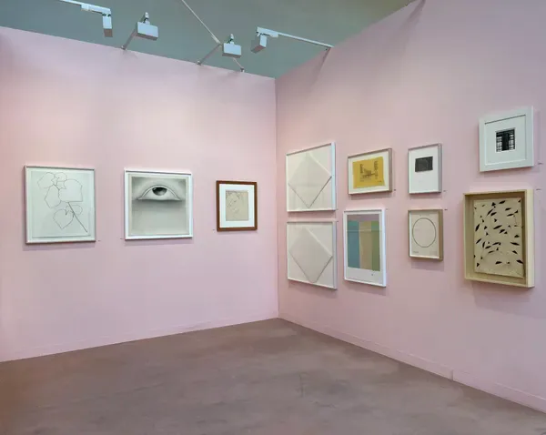 Gallery corner with pink walls displaying framed artworks of varying sizes in salon-style arrangement; light gray concrete floor.