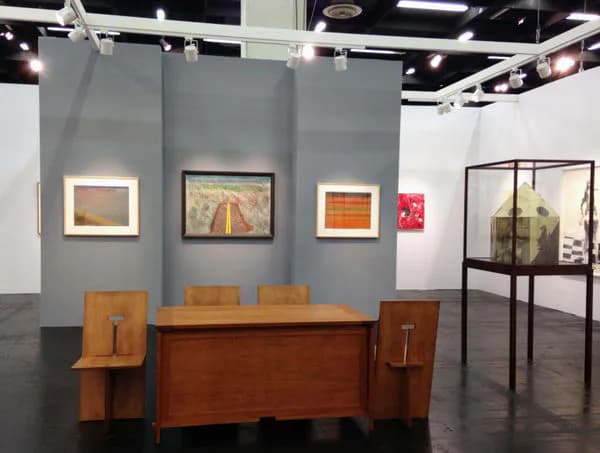 Art fair or gallery booth with gray partition walls, wooden desk, chairs, framed paintings, and display case; professional exhibition setup.