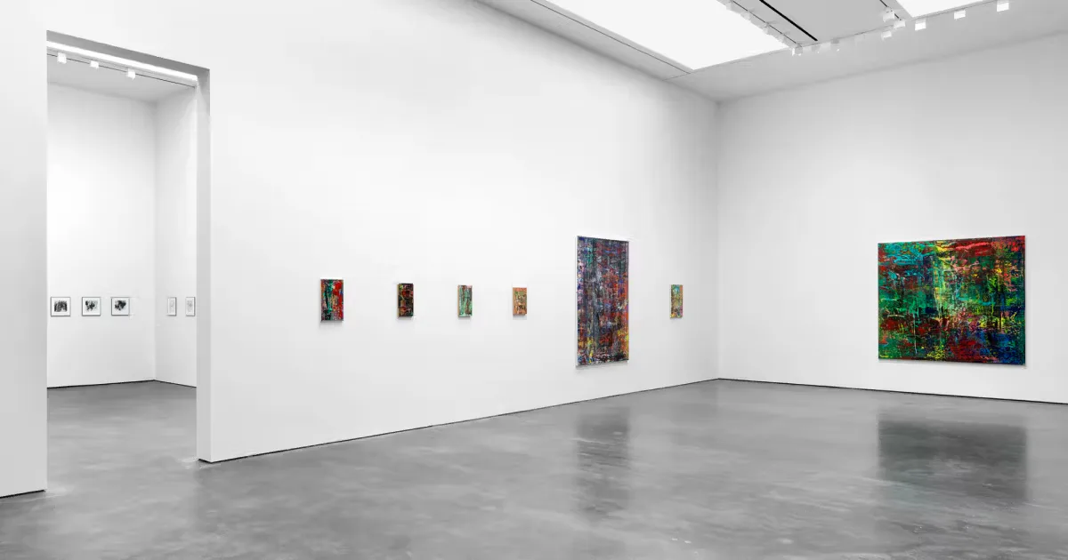 Bright gallery interior with polished gray concrete floor, white walls, and skylight. Small paintings in various sizes hung at intervals along the walls; a vibrant colorful painting visible on the right side.