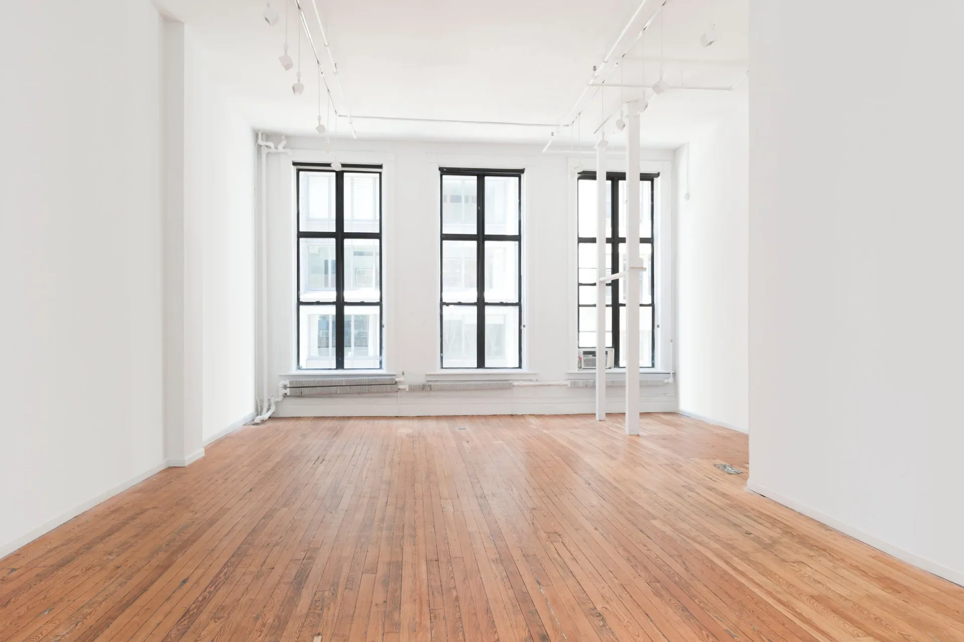 Empty gallery interior with white walls, track lighting, tall black-framed windows, and a wooden floor