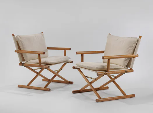 Pair of modernist wooden campaign-style chairs with cream linen cushions and X-frame folding bases, photographed against white background.