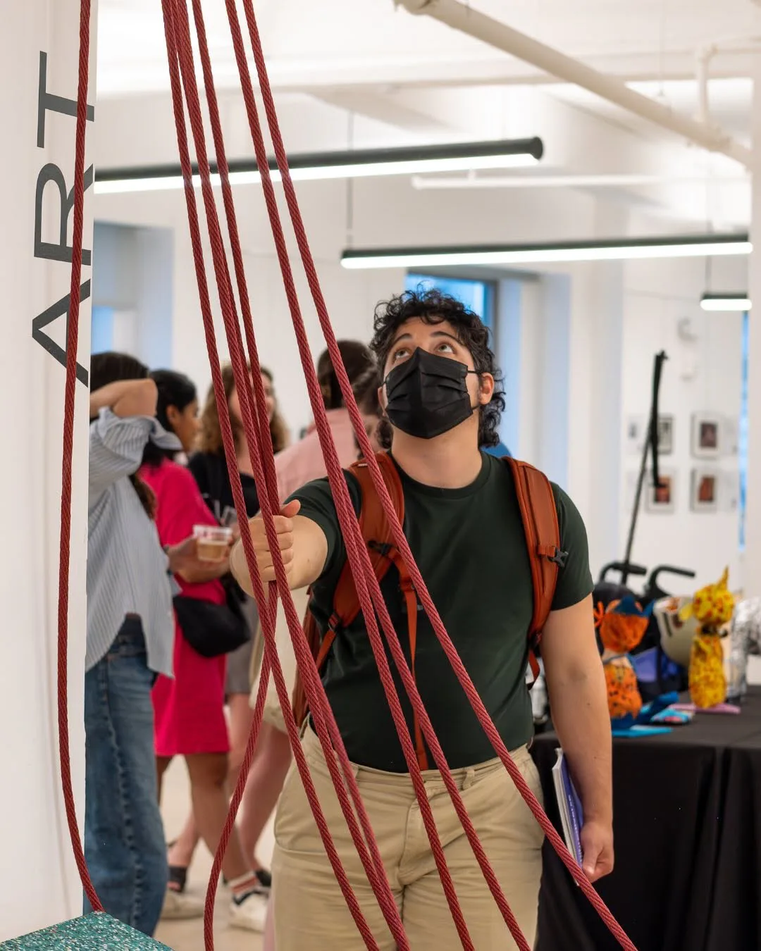 Interior gallery space with person wearing mask interacting with red rope installation; other visitors and colorful objects visible in background.