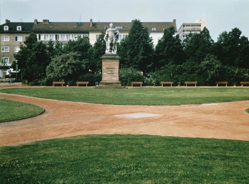 Exterior view of Dia Chelsea's plaza featuring a central statue on a beige pedestal, manicured lawn, brick pathways, and brick residential buildings in background.