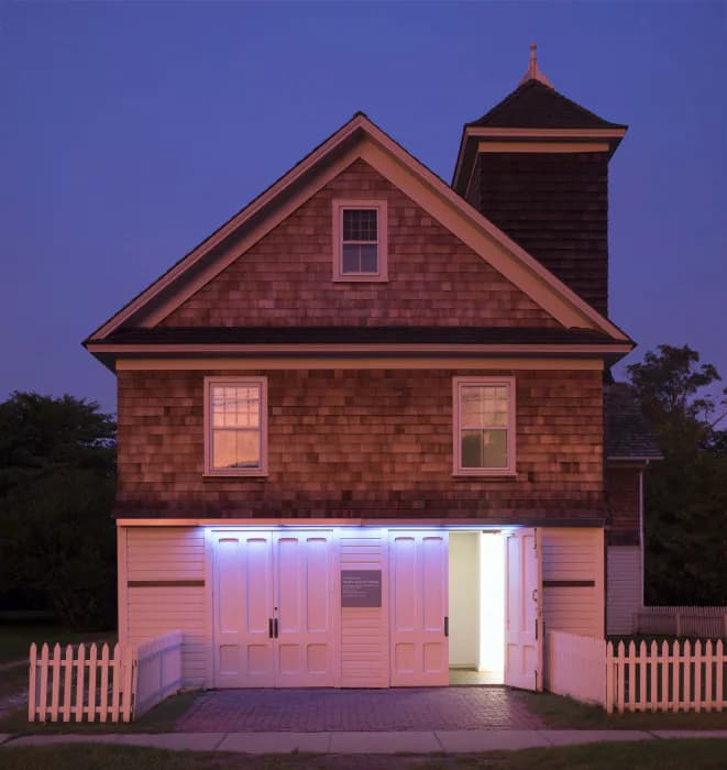 Dusk photograph of a illuminated pink cottage with white picket fence, dark shingle roof, and glowing garage doors against twilight blue sky.