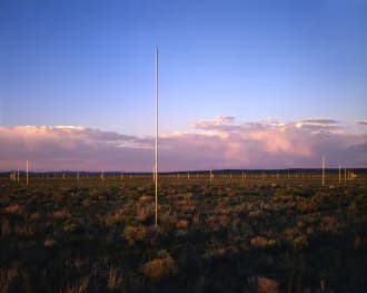 Minimalist land art: solitary vertical pole stretching across desert scrubland at dusk with golden sunset clouds and distant hills.