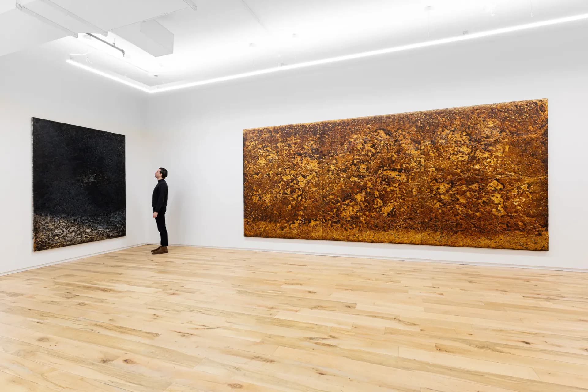 Minimalist gallery interior with wooden floor and white walls, displaying one black and one large golden-brown abstract painting.