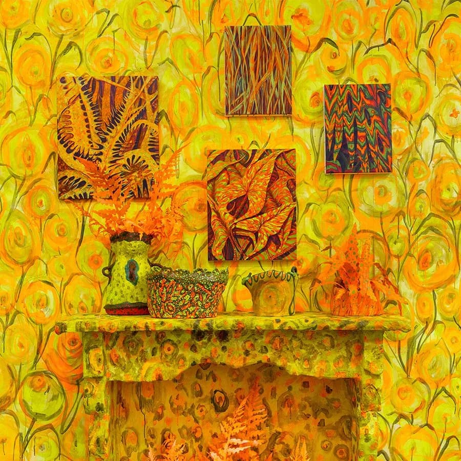 Mixed-media installation on yellow sunflower-patterned background featuring stacked shelving with various textured abstract panels in orange, brown, and multicolored patterns.