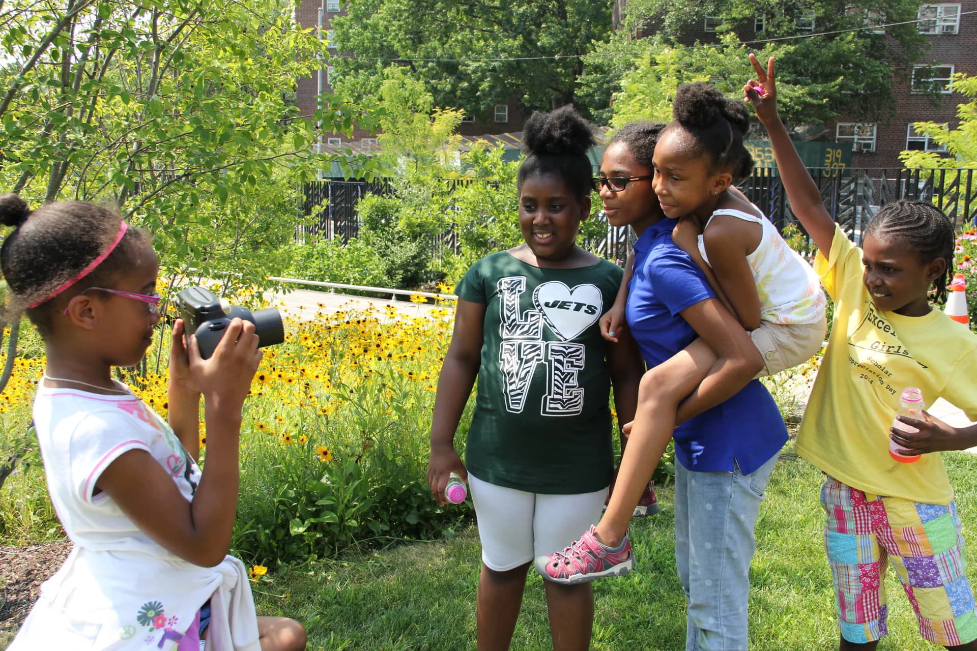 Group of young people in an outdoor urban garden with yellow flowers; one person photographs others in a documentary or participatory art context.