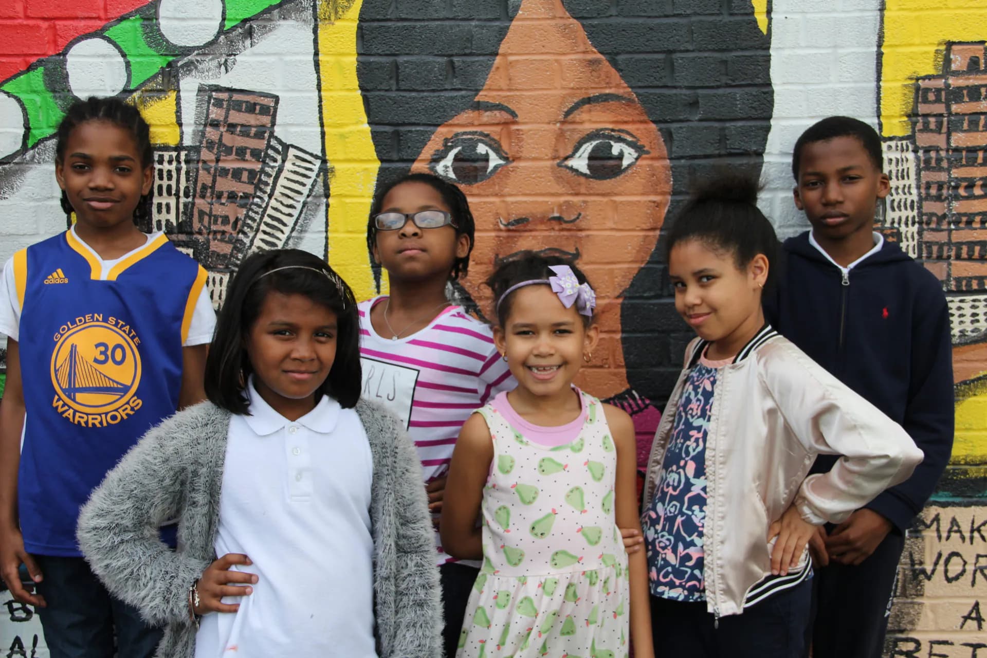 Six children posed in front of colorful mural featuring portrait of a woman's face with brown skin and bold eyes, yellow and black geometric background.