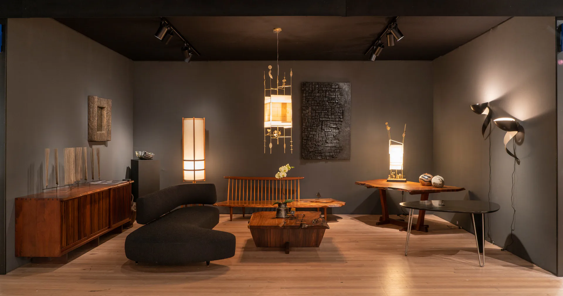 Interior gallery space at Dobrinka Salzman Gallery featuring mid-century modern furniture including wooden cabinets, tables, pendant lights, and sculptural wall art on neutral walls with wood flooring.