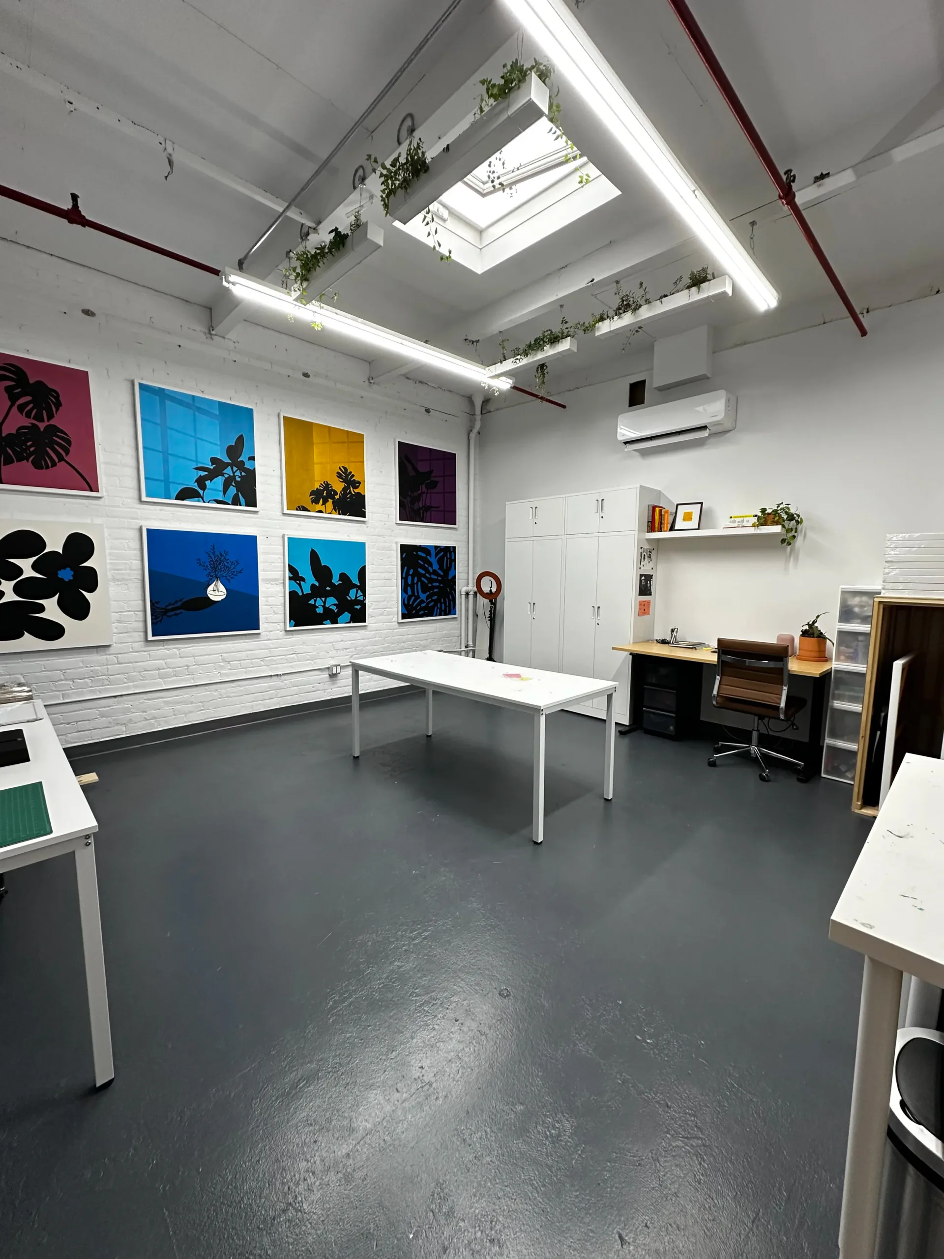 Bright studio interior with white brick walls displaying colorful framed artworks, a skylight, hanging plants, white tables, and a workspace with desk and chair