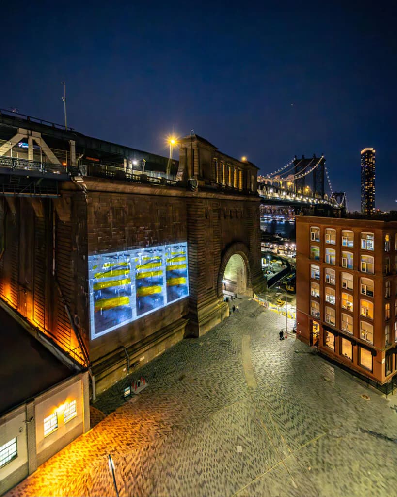 Nighttime photograph of illuminated brick buildings under a bridge in Dumbo, with digital screens and lit storefronts creating warm atmospheric lighting.