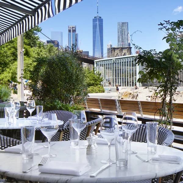 Rooftop dining terrace with round white tables, striped awning, and Manhattan skyline featuring One World Trade Center in background.