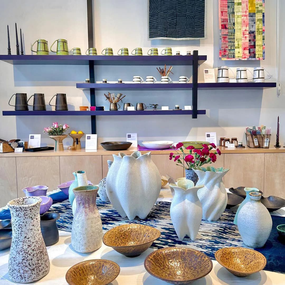 Retail or gallery interior with black floating shelves displaying white ceramics and home goods, wooden cabinetry below.