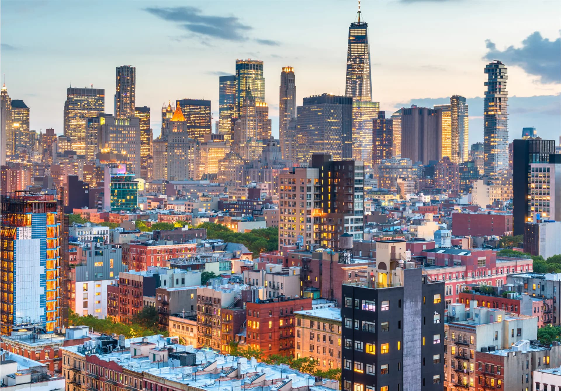 New York City skyline at dusk with illuminated skyscrapers including One World Trade Center rising above dense rows of apartment buildings