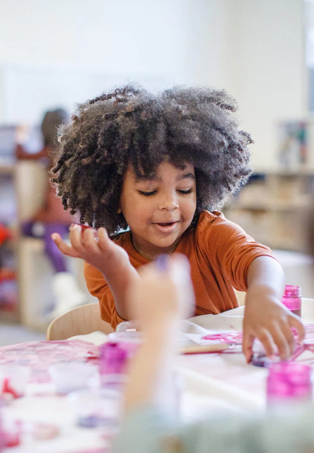 Young child with curly hair reaching over a table covered in pink paint containers during an art activity, likely at Educational Alliance