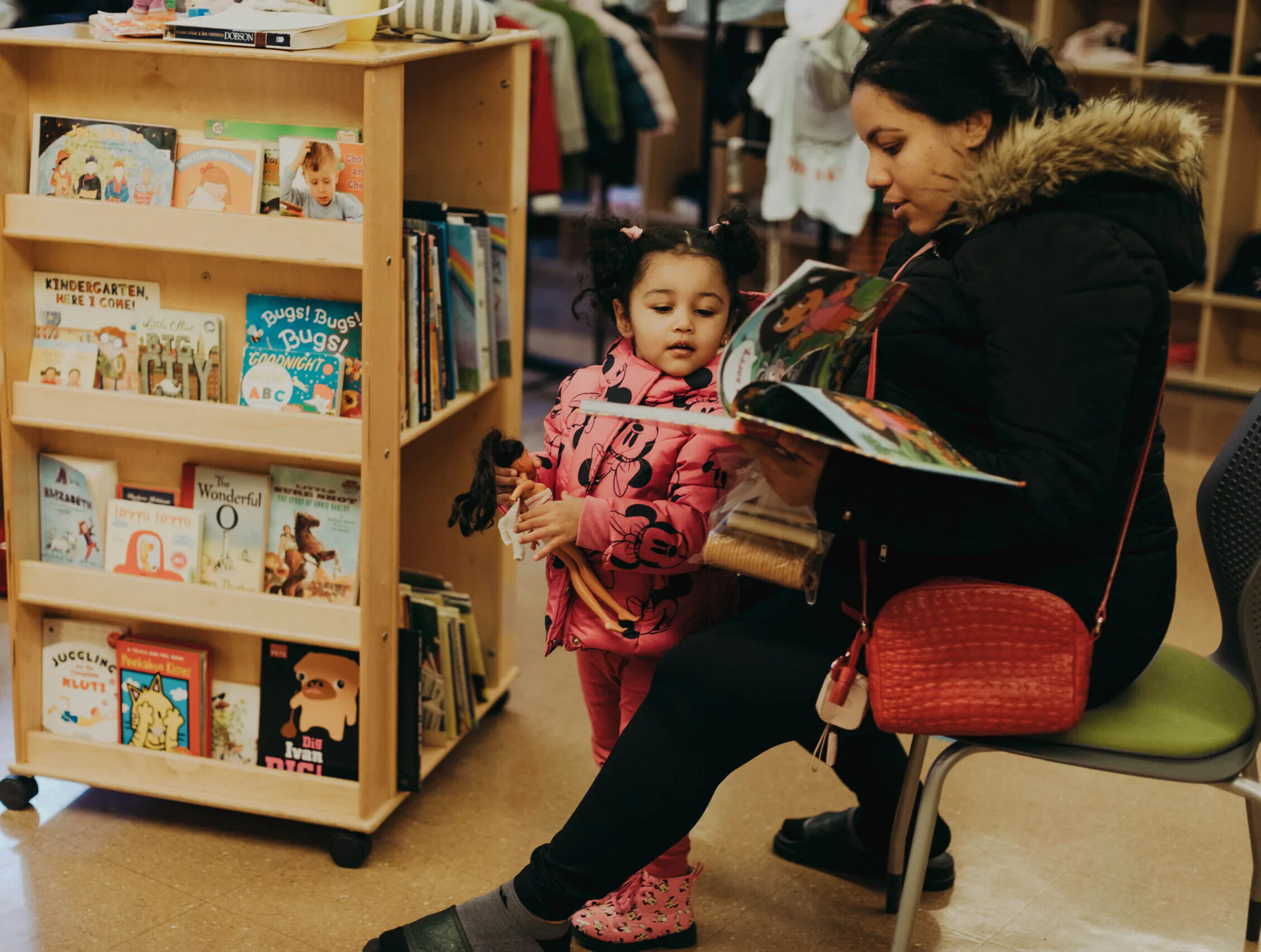 Woman in a winter coat reading a colorful picture book to a young girl beside a wooden bookshelf filled with children's books, at Educational Alliance