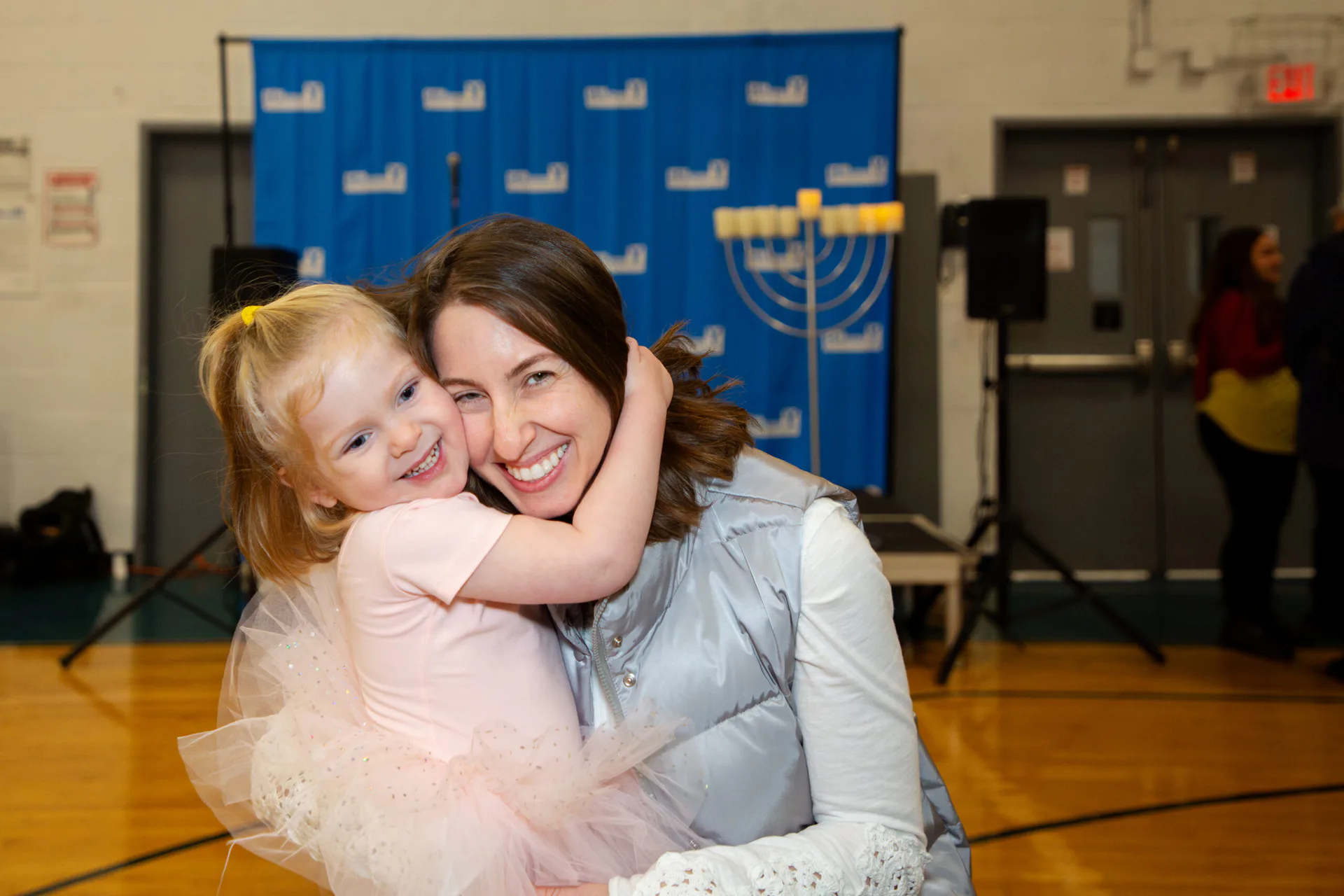 Woman smiling and hugging a little girl in a pink tutu in front of a blue backdrop with a menorah, at an Educational Alliance event