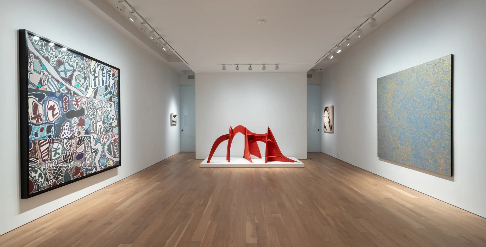 Modern gallery interior with polished wood floors, white walls, track lighting, colorful abstract paintings on left and right walls, and a red sculptural installation in center.