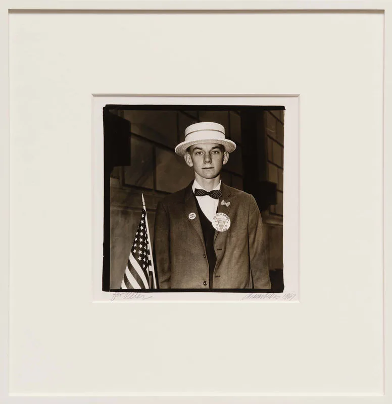 Black and white vintage photograph of a man wearing a straw hat, bow tie, and jacket with an American flag pin, mounted on cream mat.