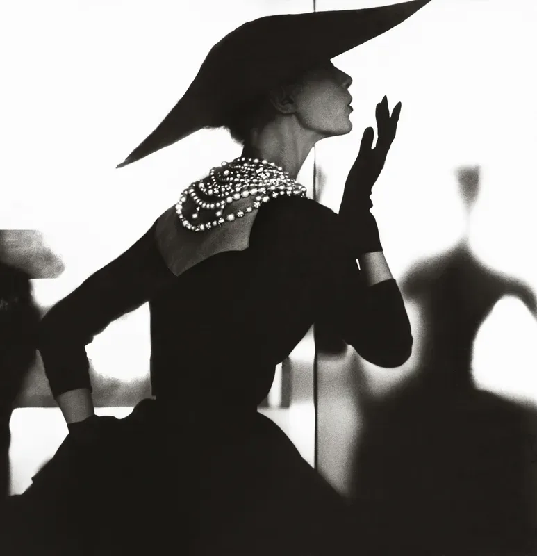 Black and white silhouette photograph of a woman in profile wearing a dramatic pointed hat and pearl necklace, hands raised expressively.