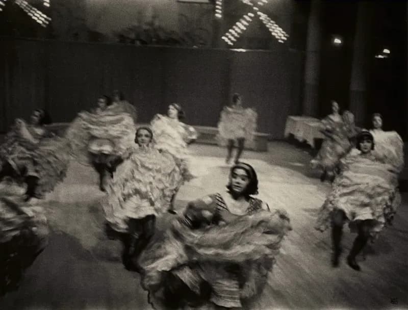 Blurred black and white photograph of performers in voluminous period costumes dancing or moving on a theatrical stage.