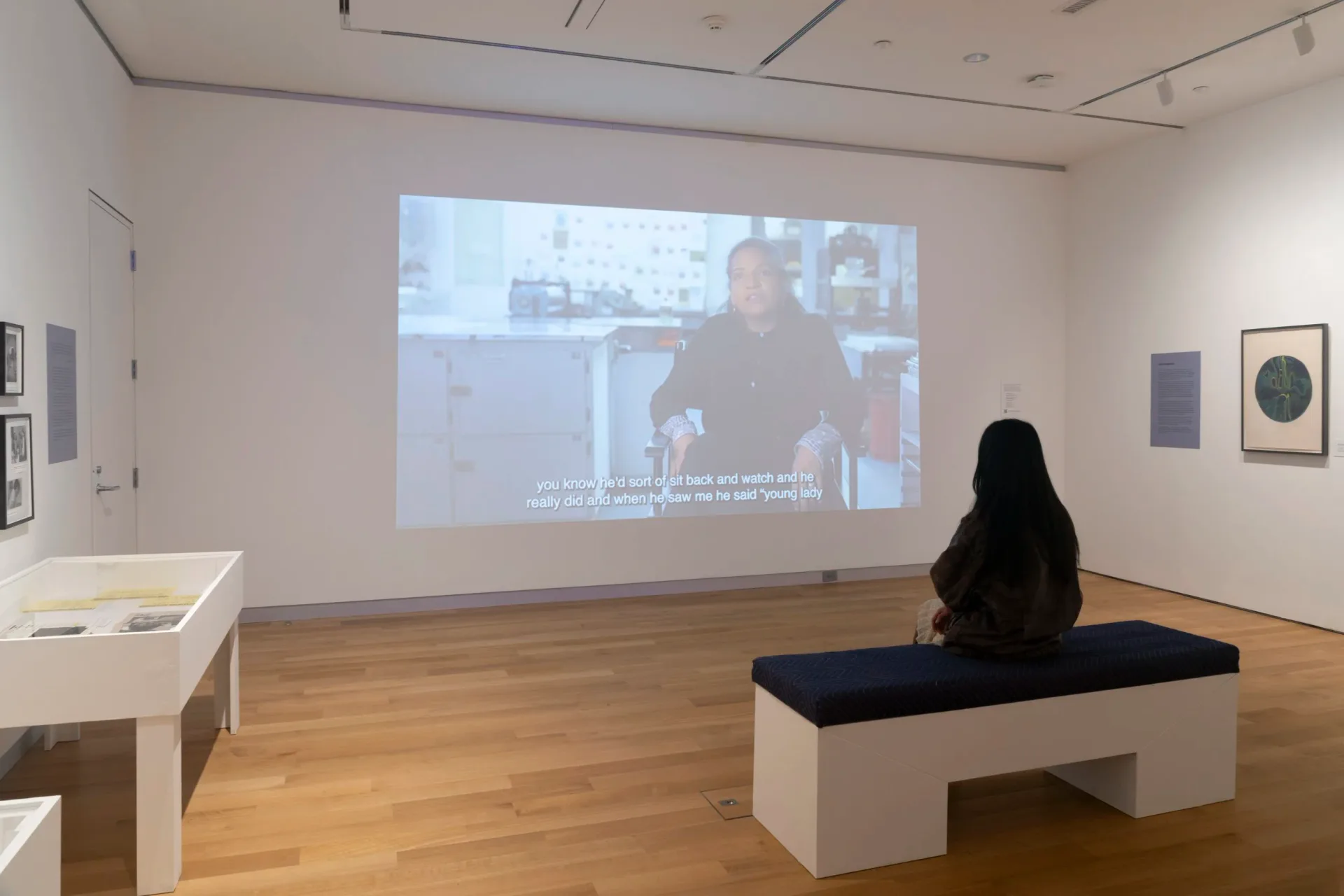 Gallery interior with wooden floors, white walls, and mounted artworks; a person sits on a bench watching a projected video.