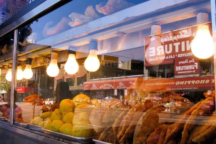 Warm-toned photograph of a shop window display with hanging produce, warm lighting, and reversed signage visible through glass.