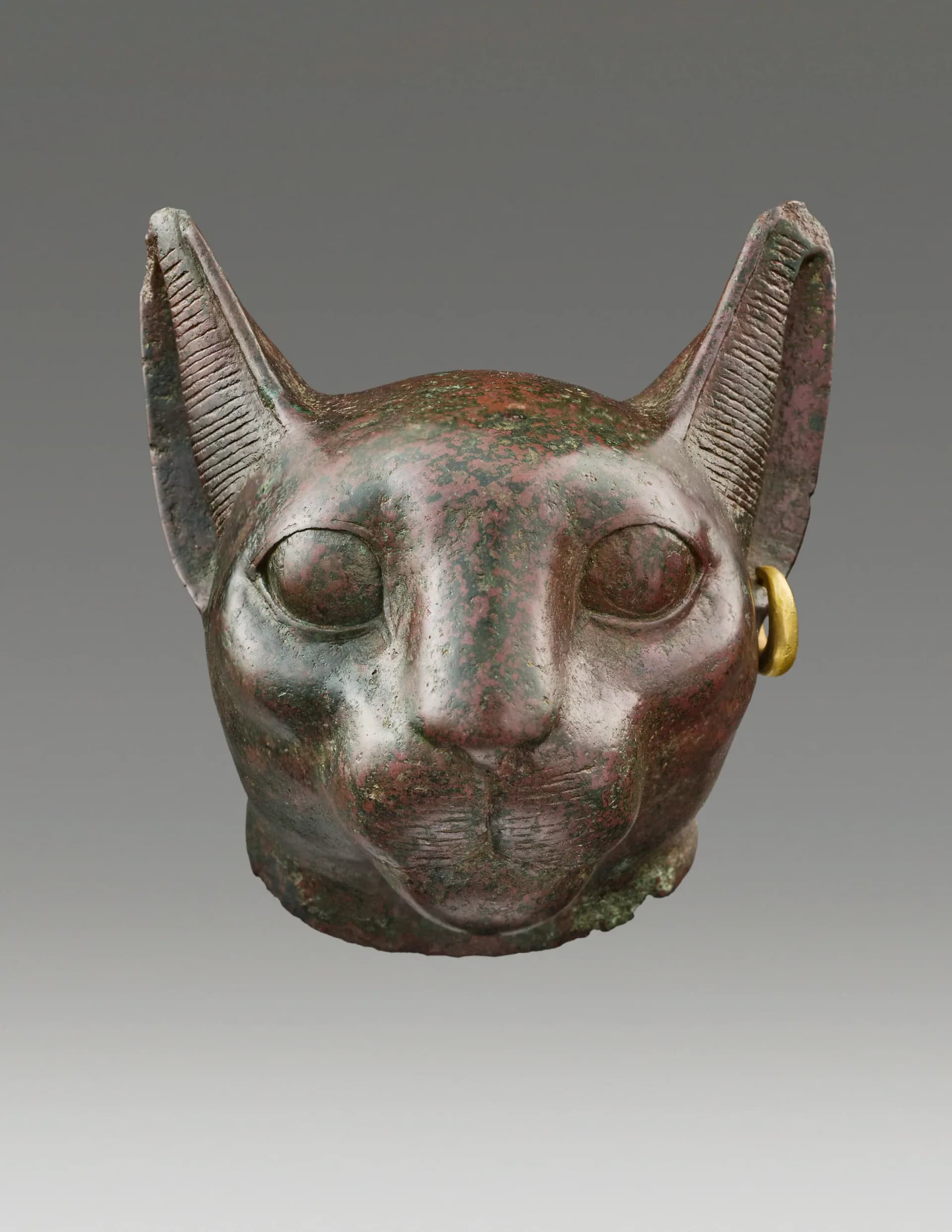 Front-facing bronze cat head with protruding ears and gold hoop earring, patina showing green and brown tones.