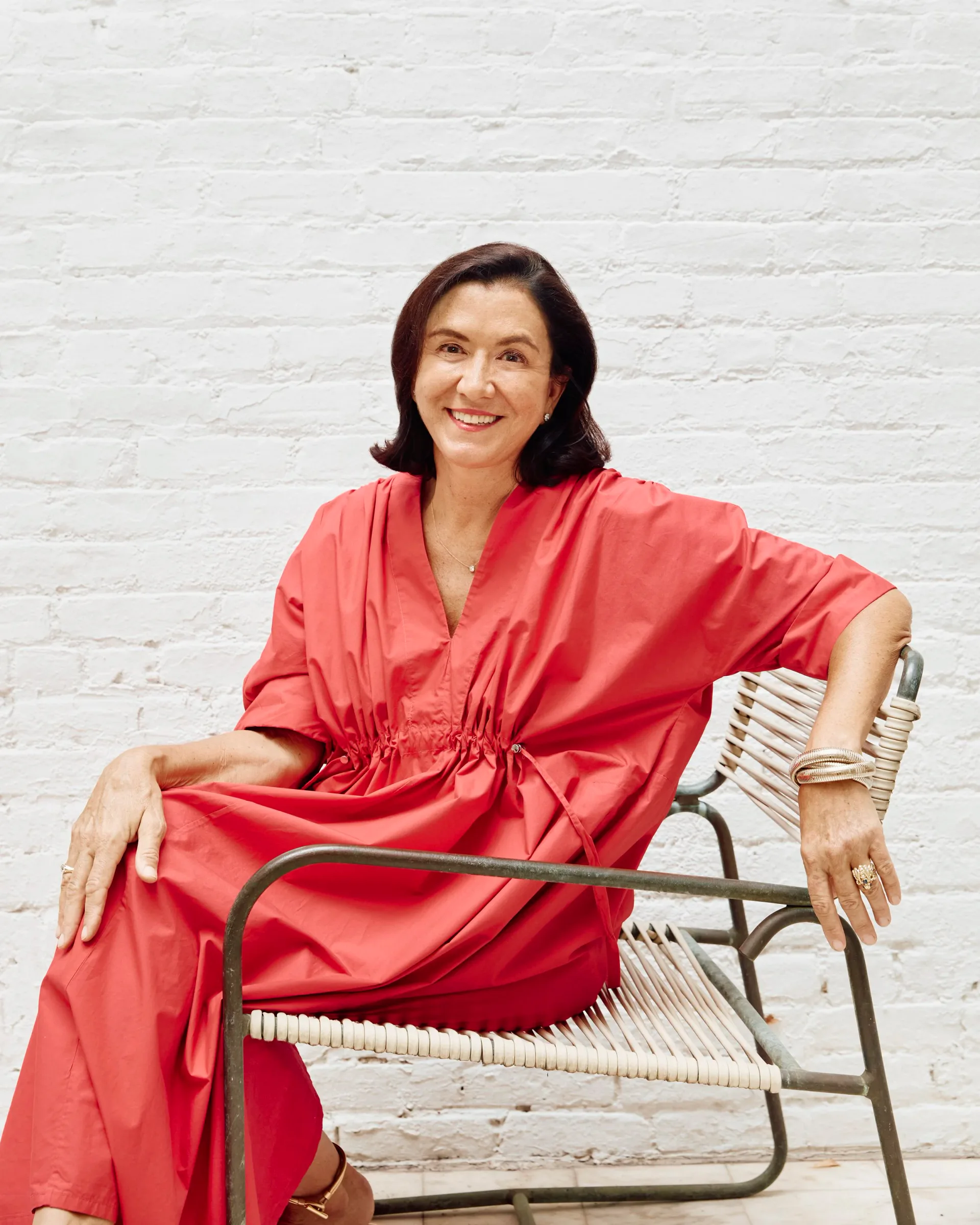Woman in bright coral wrap dress seated on modern metal chair against white brick wall, smiling at camera.
