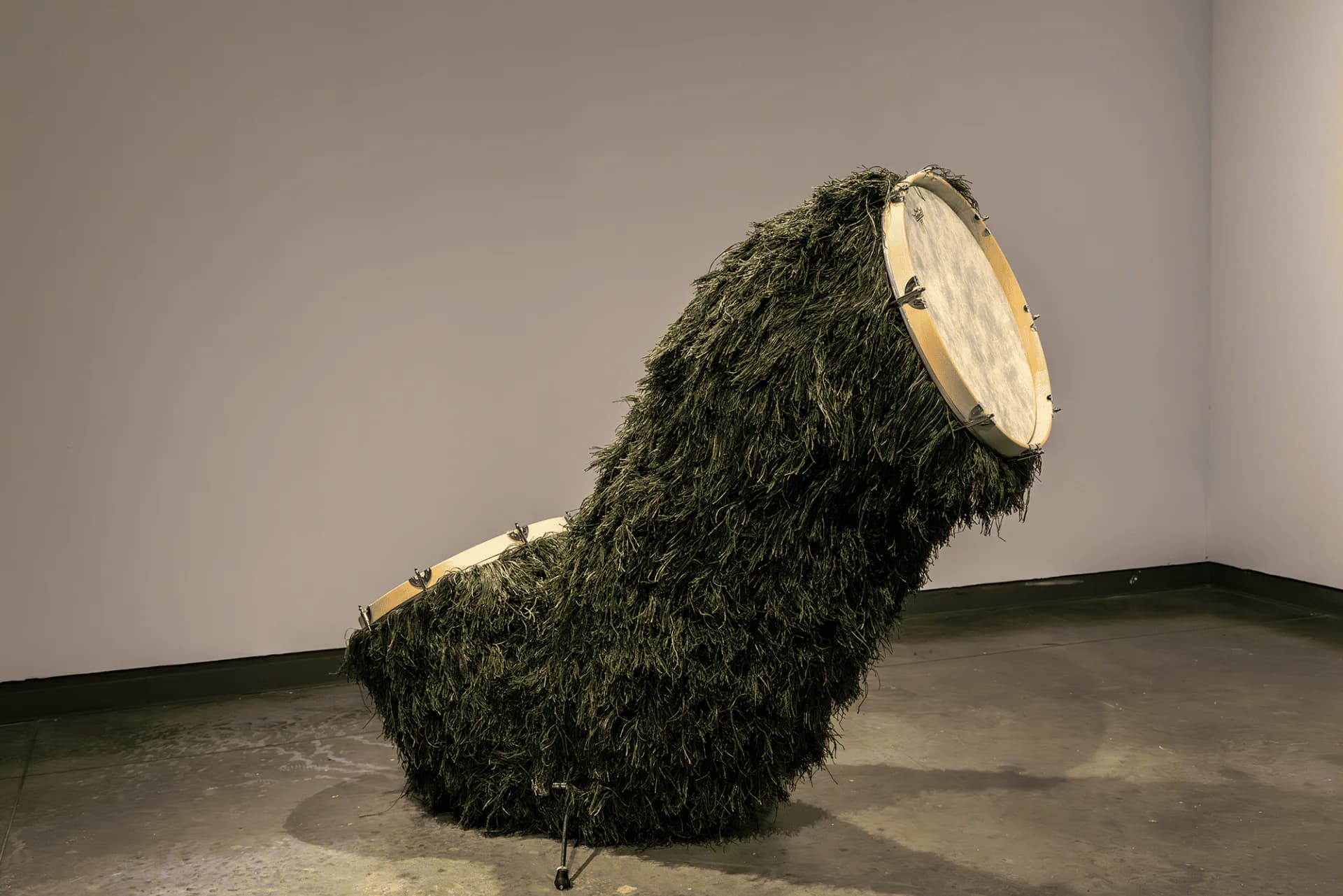 Large dark moss-covered sculptural form with a pale circular opening, installed on a concrete gallery floor.