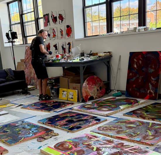 Artist working at table in bright studio space with large windows; colorful paintings and works displayed on floor and walls.